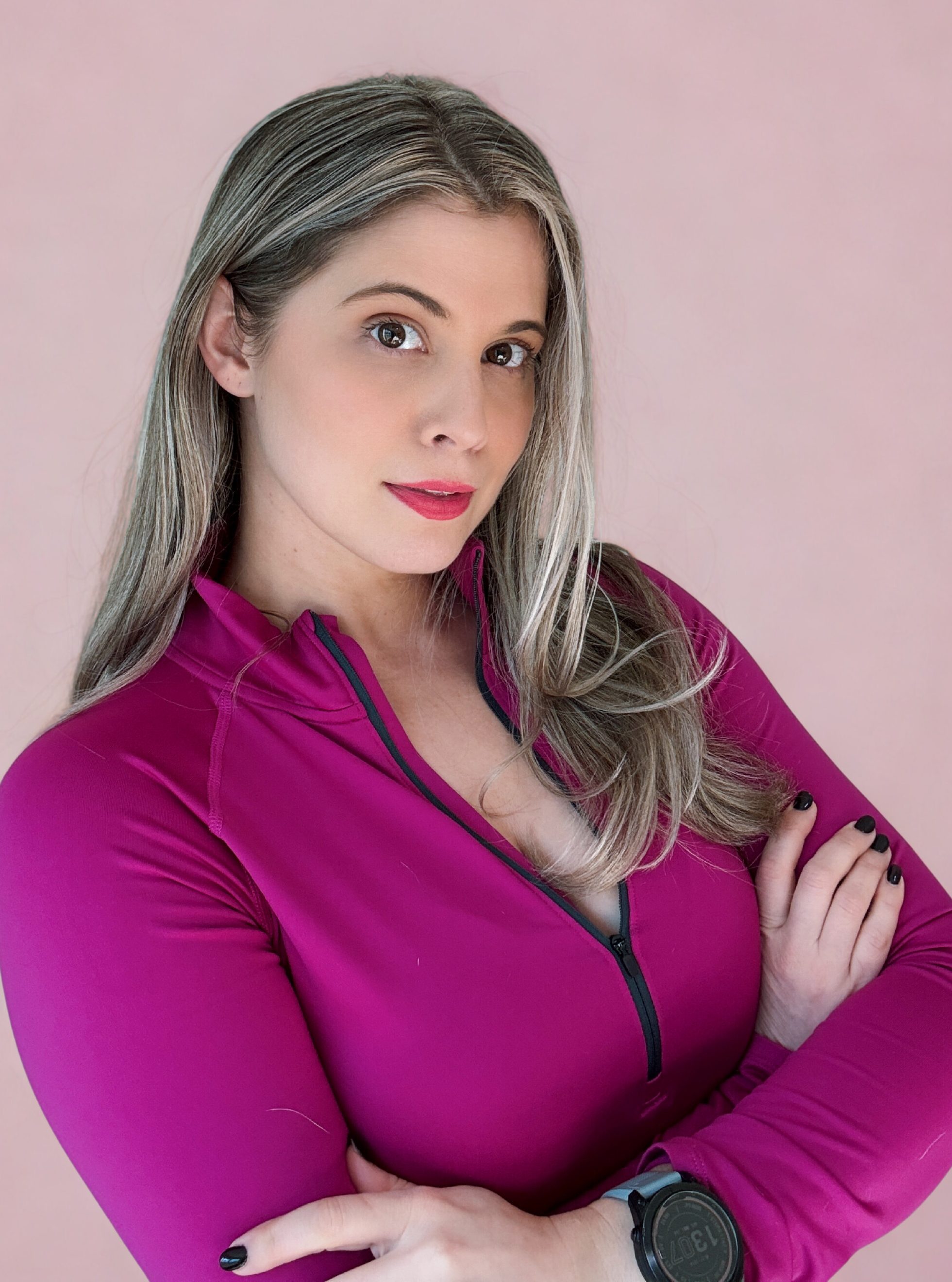 A woman with long, light brown hair poses confidently with her arms crossed, wearing a magenta athletic top against a pink background.